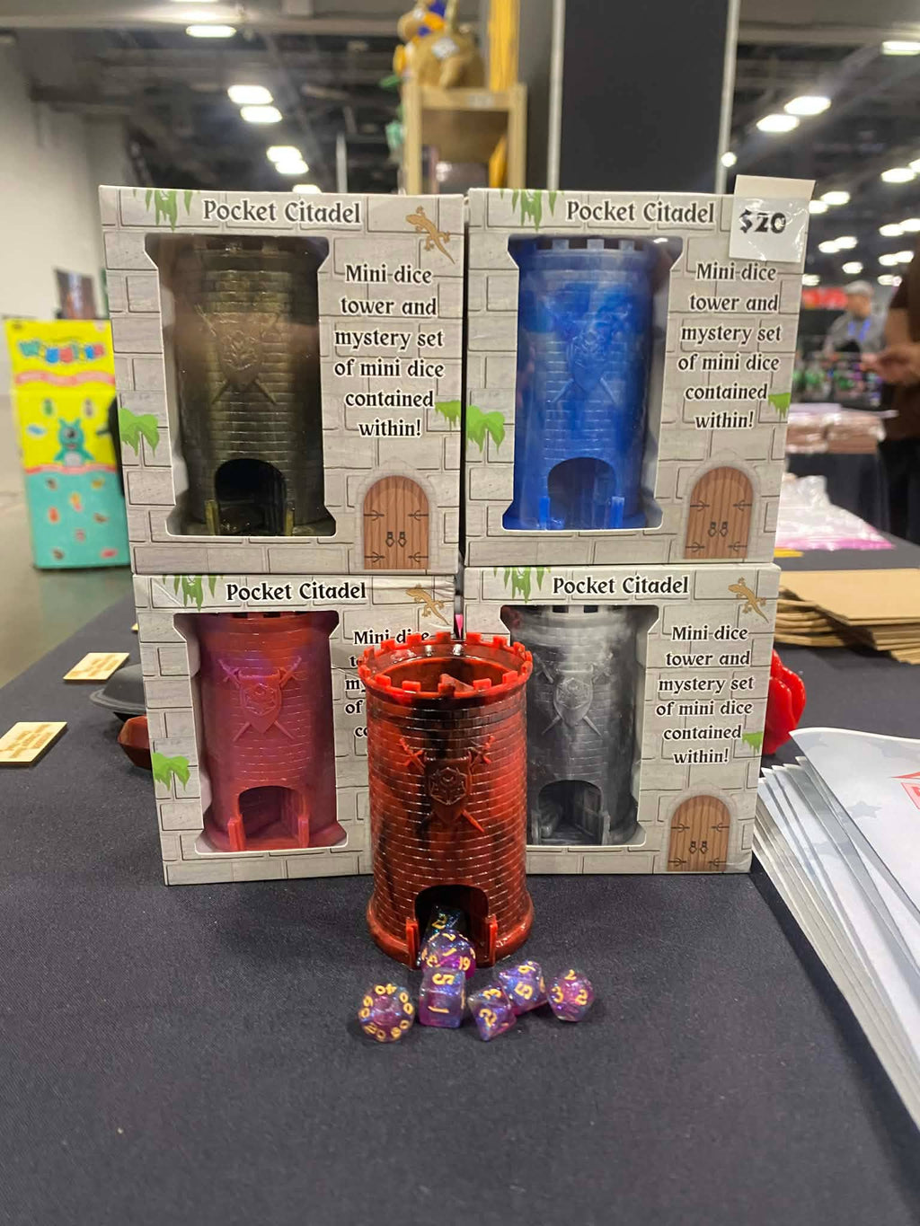 Four boxed 'Pocket Citadel' mini dice towers and mystery sets arranged on a table, with one red tower and several purple polyhedral dice placed in front. The boxes show different color options: dark green, blue, red, and gray.
